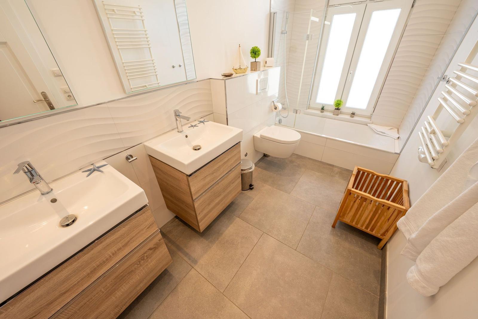 Badezimmer mit Dusche, Waschbecken und Toilette. Holzunterschränke und Fenster.