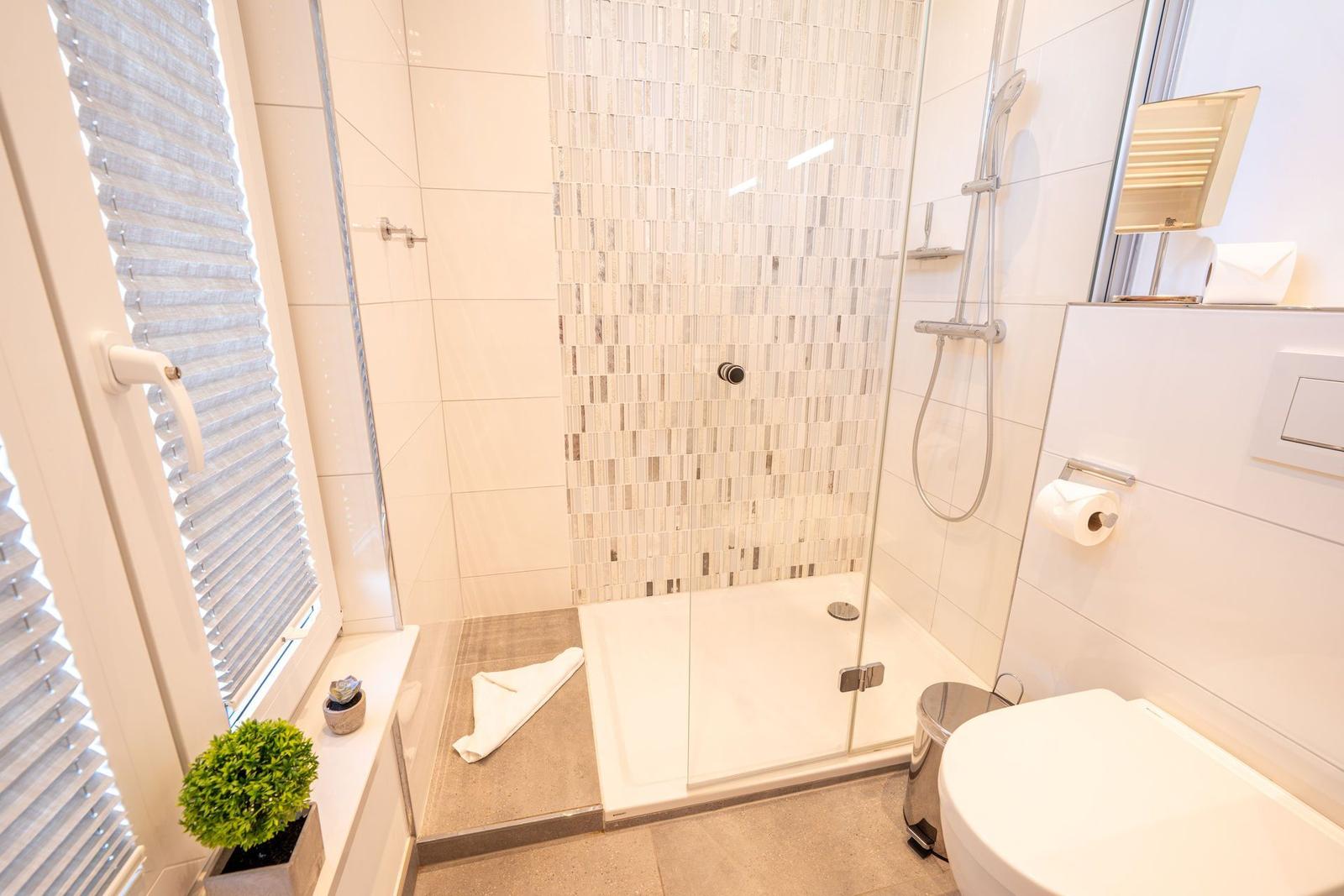 Badezimmer mit Dusche, Toilette und Fenster mit Jalousie.