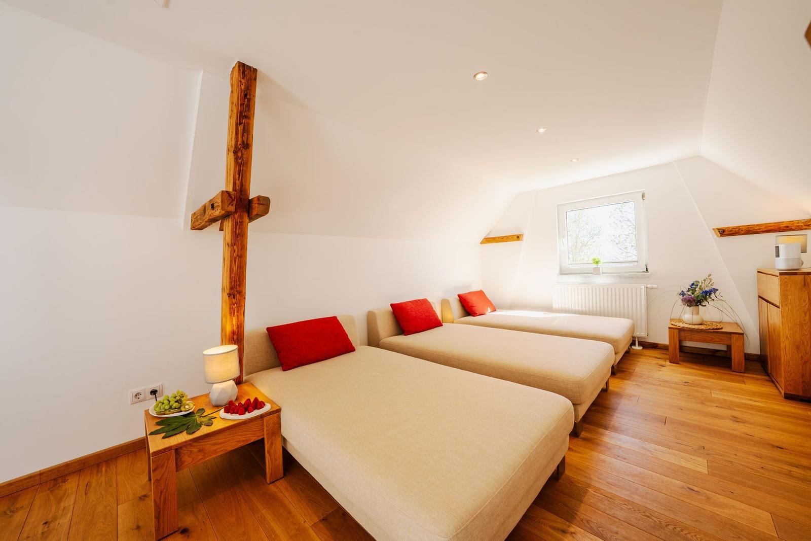 Zimmer mit zwei Betten, Holzboden und Fenster. Holzkreuz an der Wand.