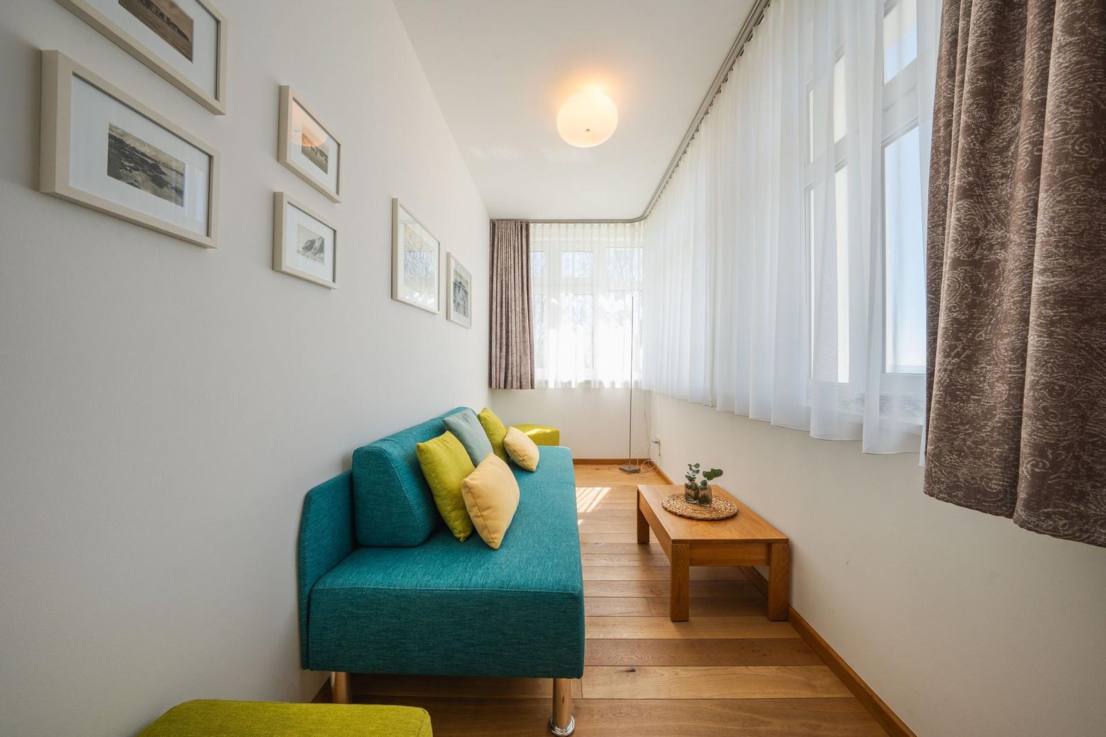 Ein Wohnzimmer mit blauem Sofa, Holzboden und Fenstern mit weißen und braunen Vorhängen.