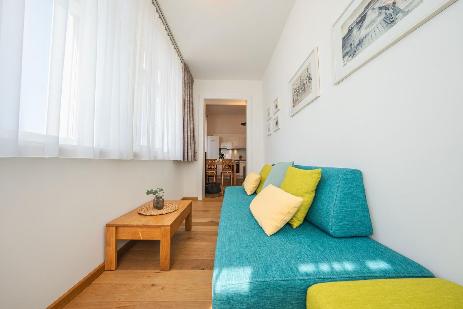 Ein Wohnzimmer mit blauem Sofa, gelben Kissen und Holztisch. Durchgang zur Küche.