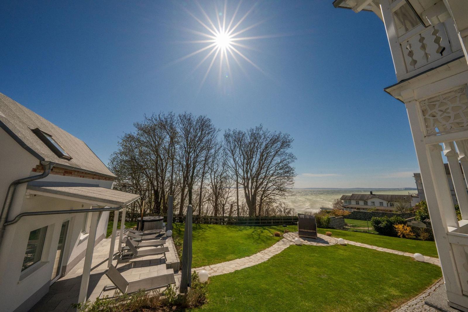 Haus mit Terrasse, Garten und Blick auf Wasser unter blauem Himmel.