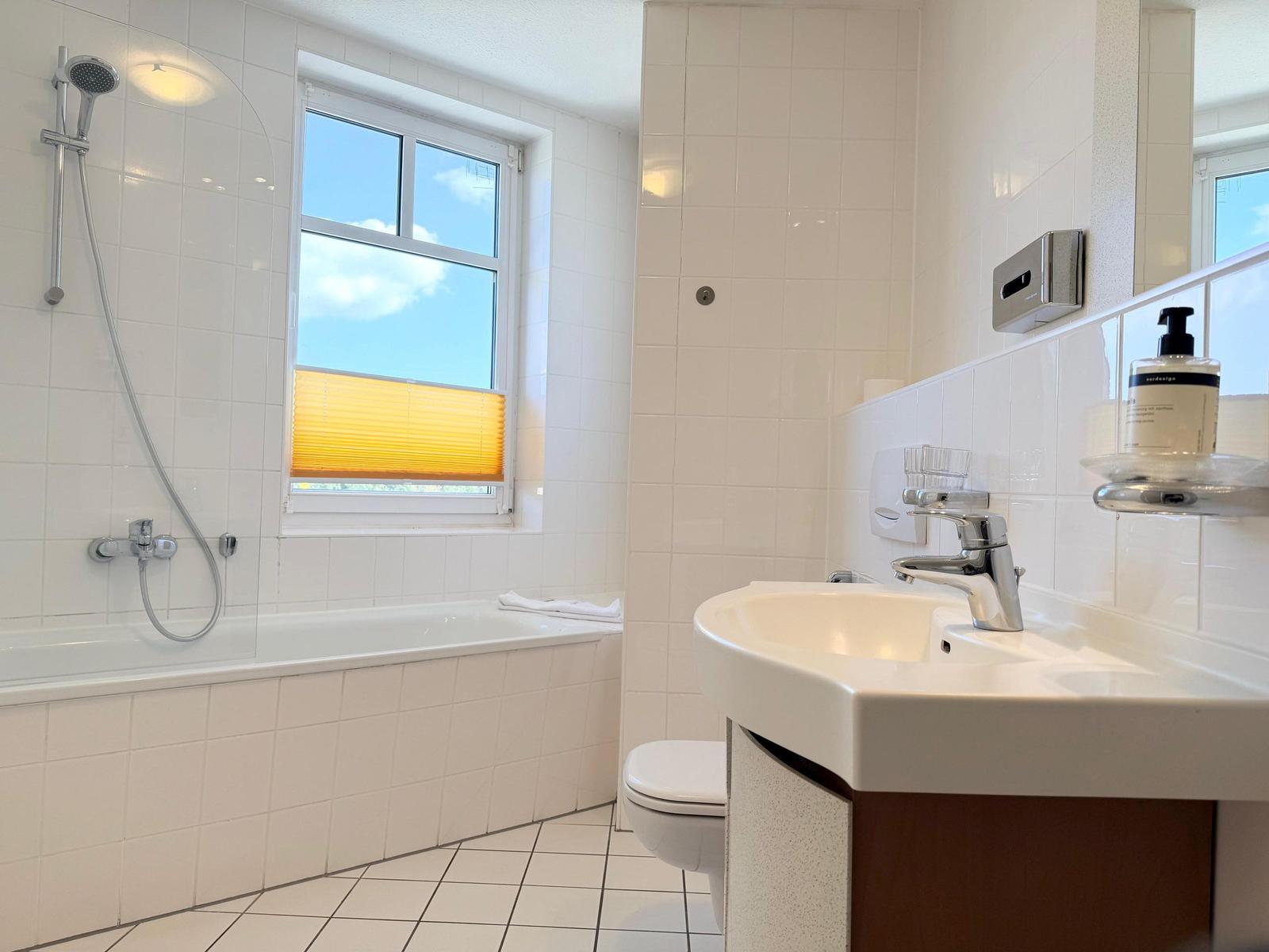 Badezimmer mit Badewanne, Dusche, Waschbecken und Toilette. Fenster mit Jalousie.