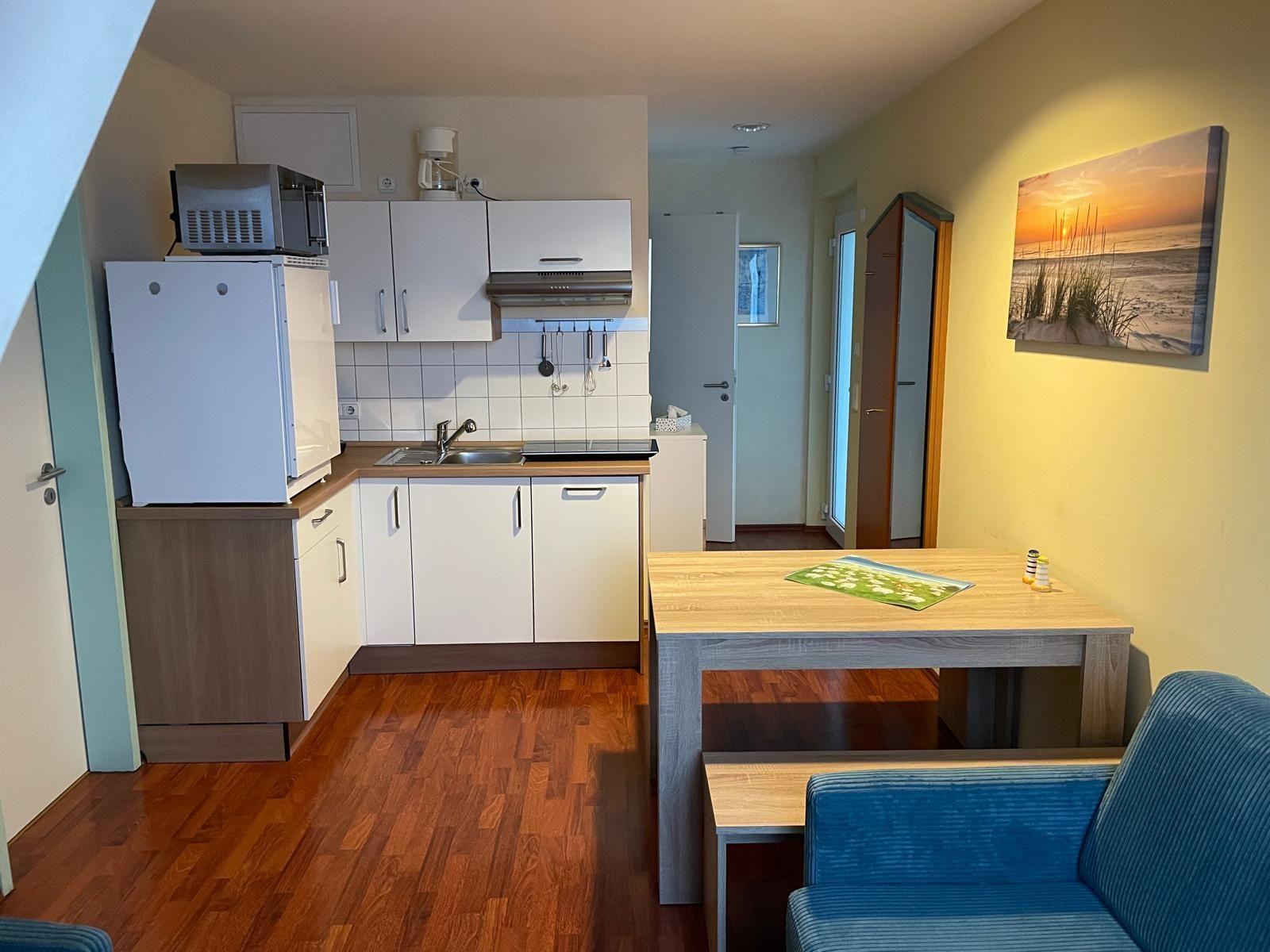 Küche mit Arbeitsplatte, Spüle und Kühlgerät. Wohnbereich mit Tisch, Sofa und Wandbild.