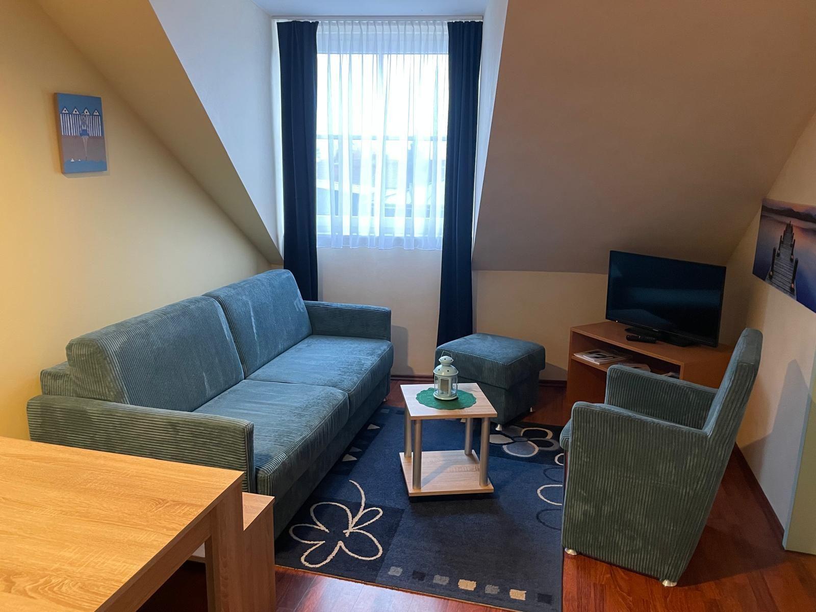 Wohnzimmer mit Sofa, Sessel, Fernseher und Fenster mit Vorhängen.