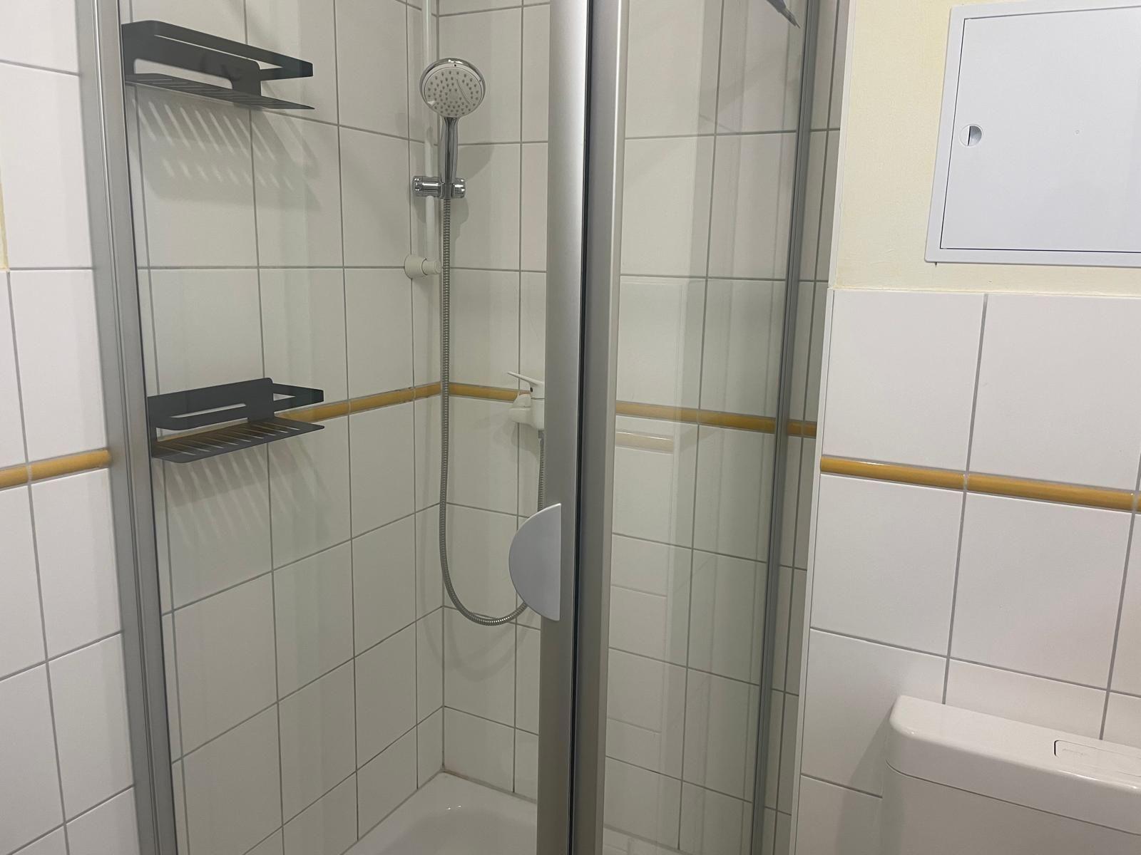 Dusche mit Glaswand, Duschbrause und zwei schwarzen Halterungen an weißen Fliesen.