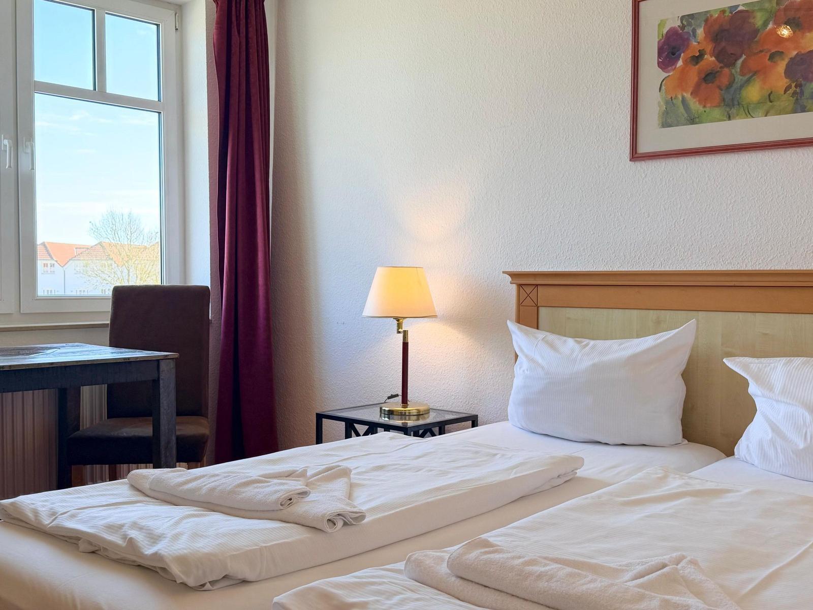 Zimmer mit Doppelbett, Nachttisch, Lampe, Fenster und Wandbild.