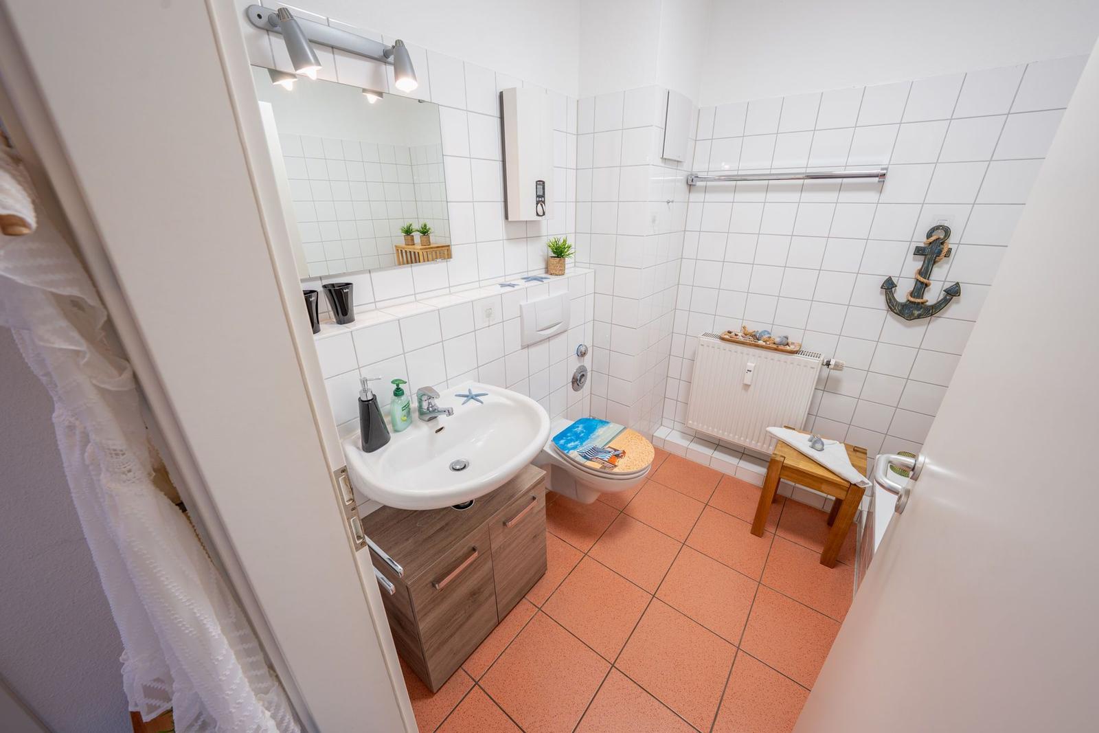 Badezimmer mit Waschbecken, Toilette und Dusche. Weiße Fliesen, orangefarbener Boden und Anker-Dekoration.