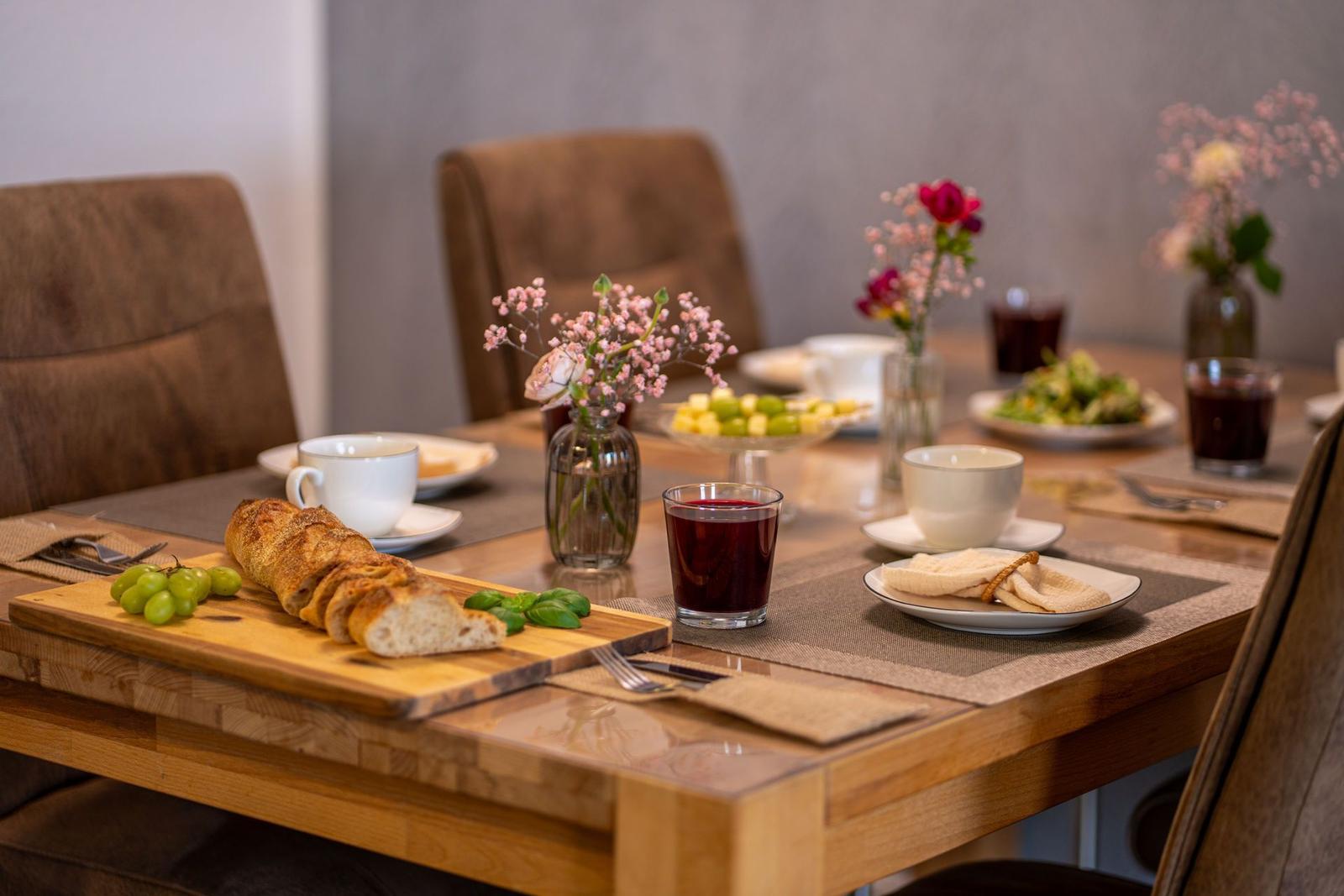 Ein gedeckter Holztisch mit Brot, Obst und Getränken. Blumenarrangements und Teller sind sichtbar.