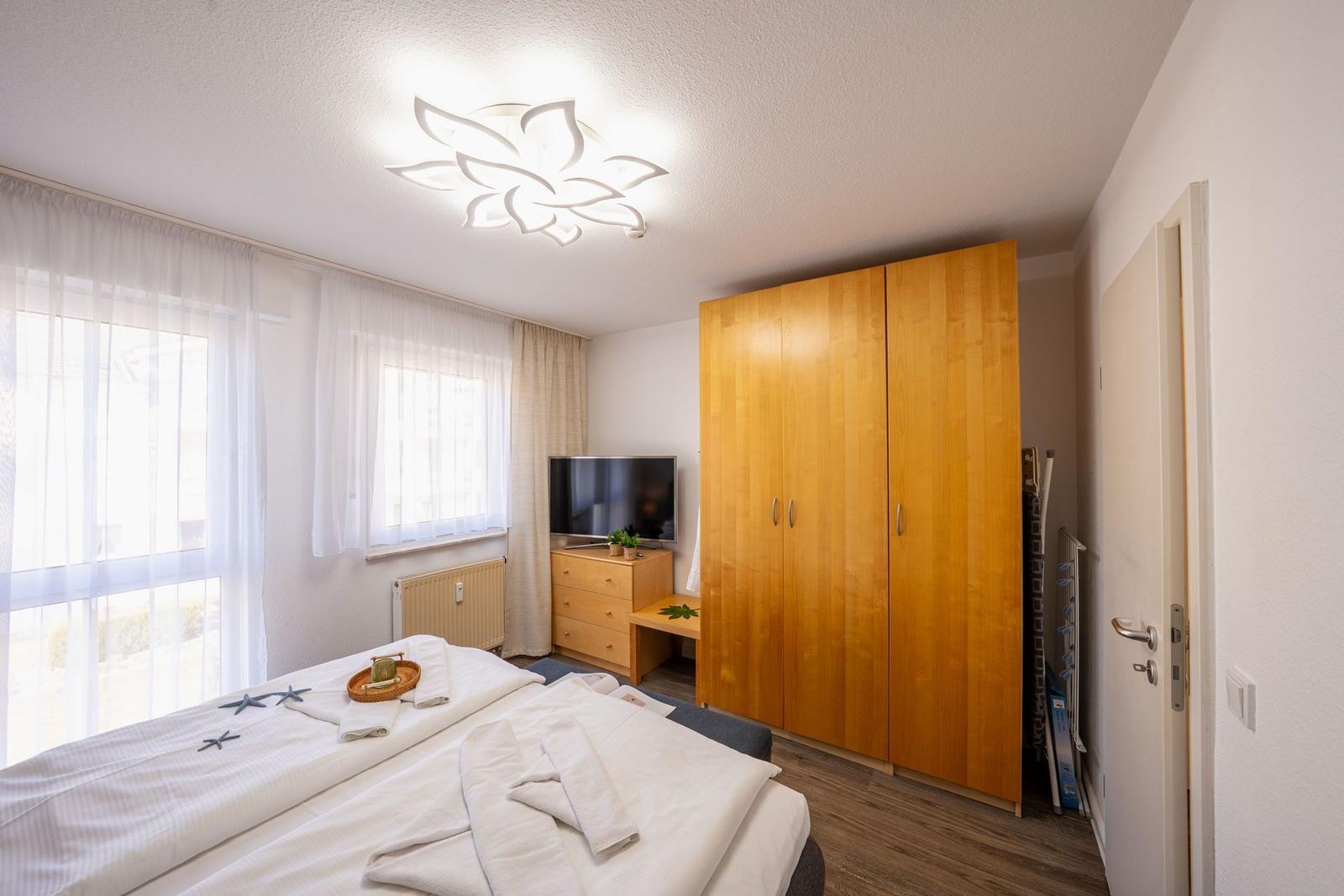 Schlafzimmer mit Bett, Kleiderschrank und TV. Fenster mit Vorhängen und Deckenleuchte.