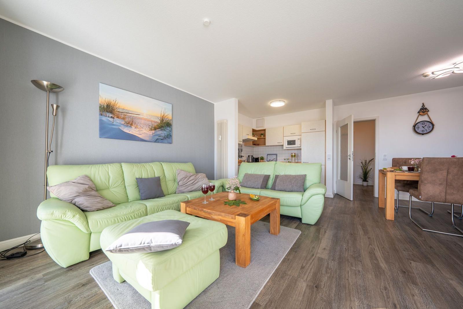 Wohnbereich mit grünen Sofas, Holztisch und Küchenbereich im Hintergrund.