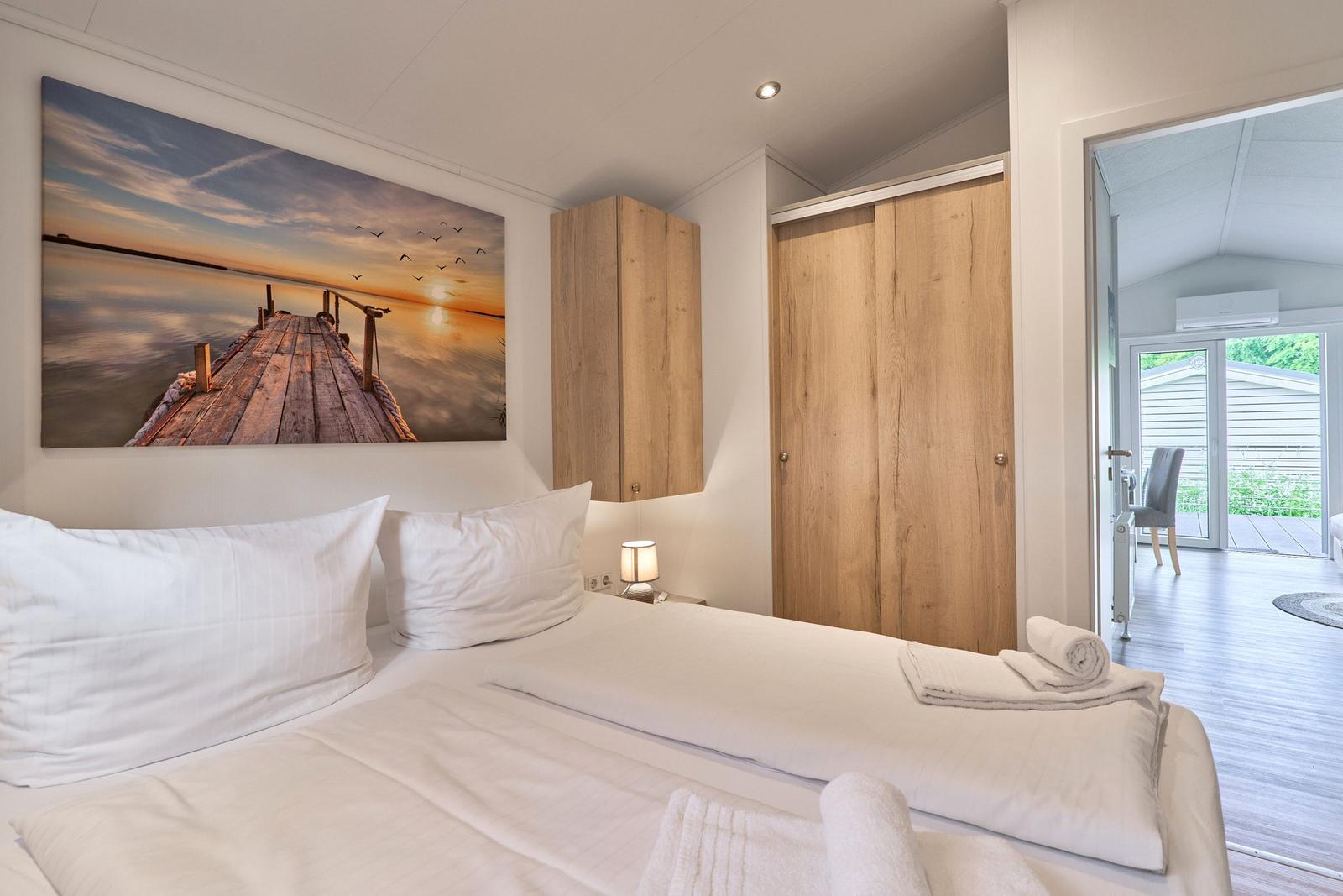 Schlafzimmer mit Bett, Wandbild und Holzschränken. Blick in Wohnbereich mit Terrasse.