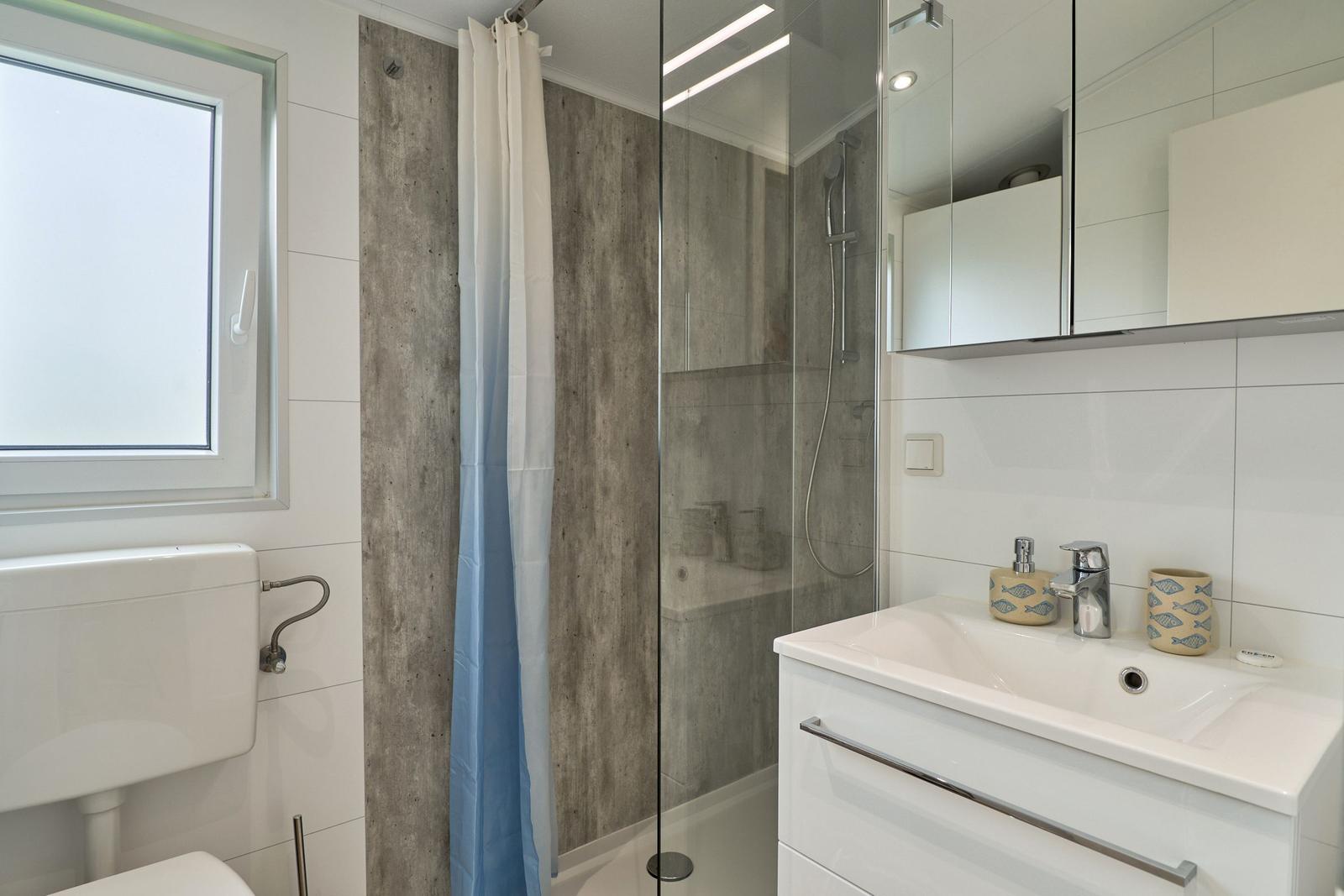 Badezimmer mit Dusche, Waschbecken und Toilette. Fenster und Spiegelschrank sichtbar.