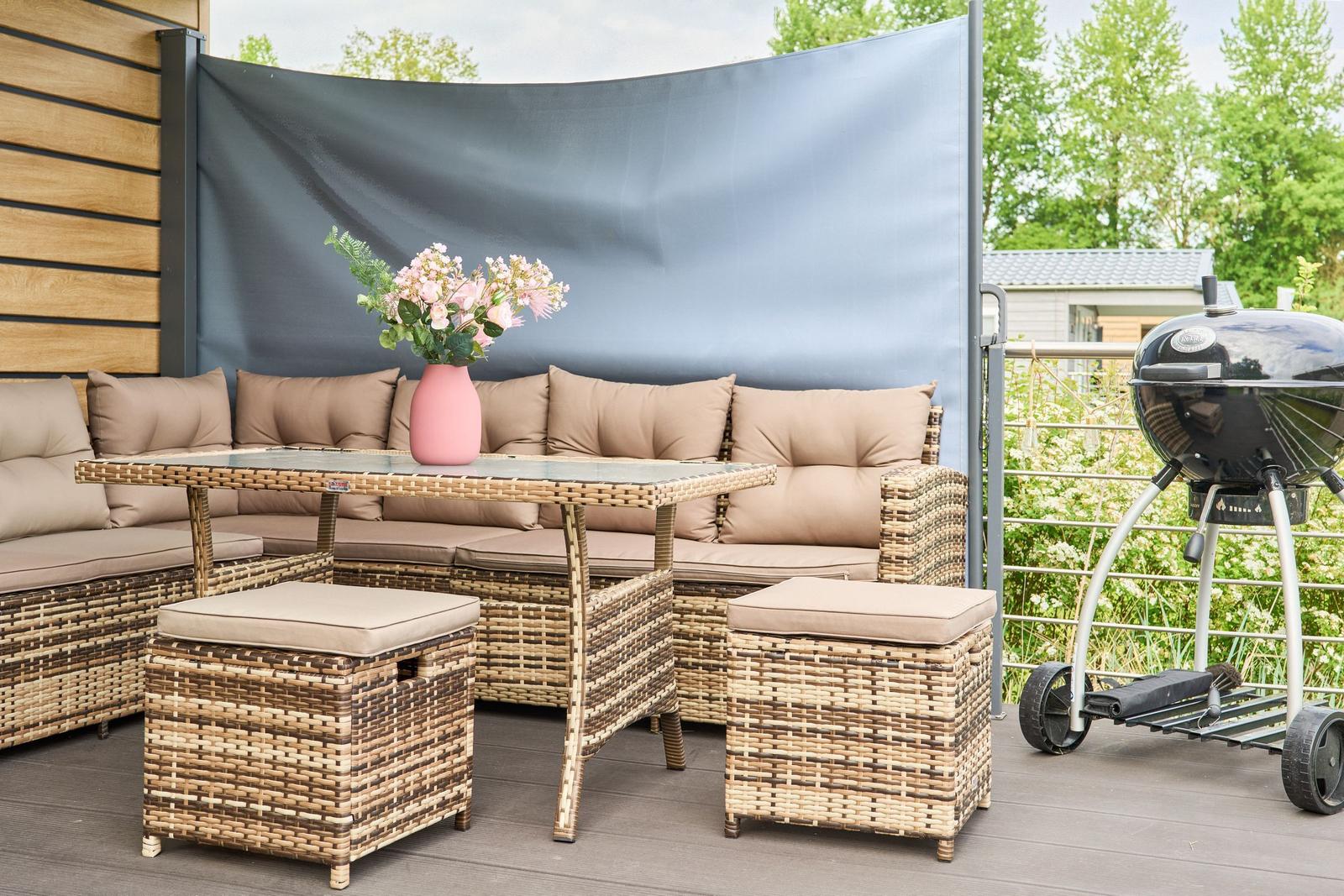 Terrasse mit Sitzgruppe, Tisch, Blumen und Grill. Hintergrund: Holzwand und Bäume.