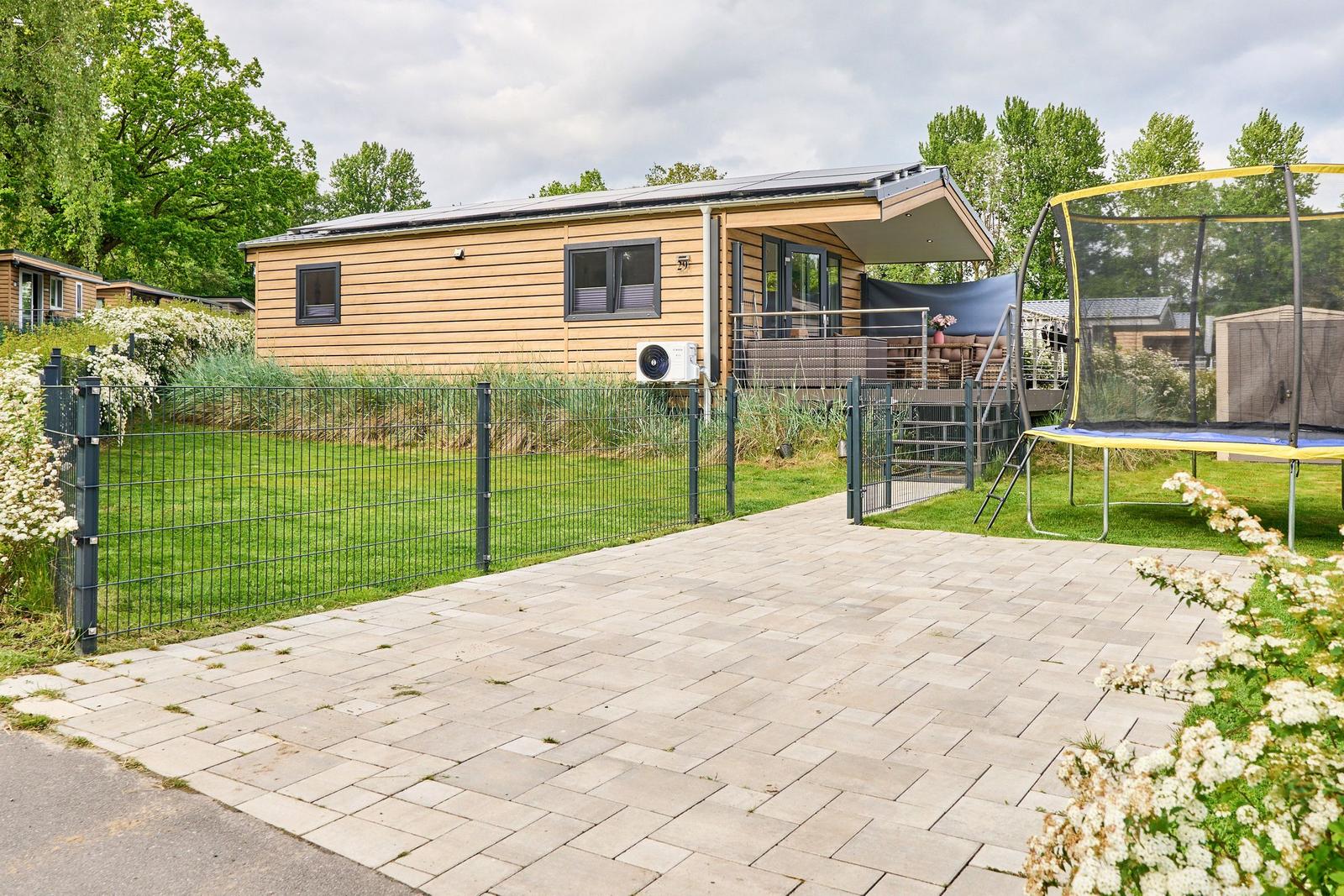 Moderne Holzhütte mit Terrasse, Trampolin und gepflastertem Hof. Umgeben von Grün und Bäumen.
