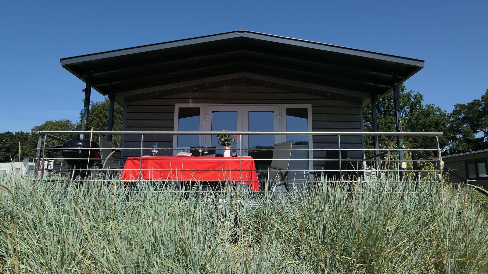 Terrasse mit Tisch, Stühlen und Grill unter schwarzem Dach. Roter Tischdecke und Pflanzen sichtbar.