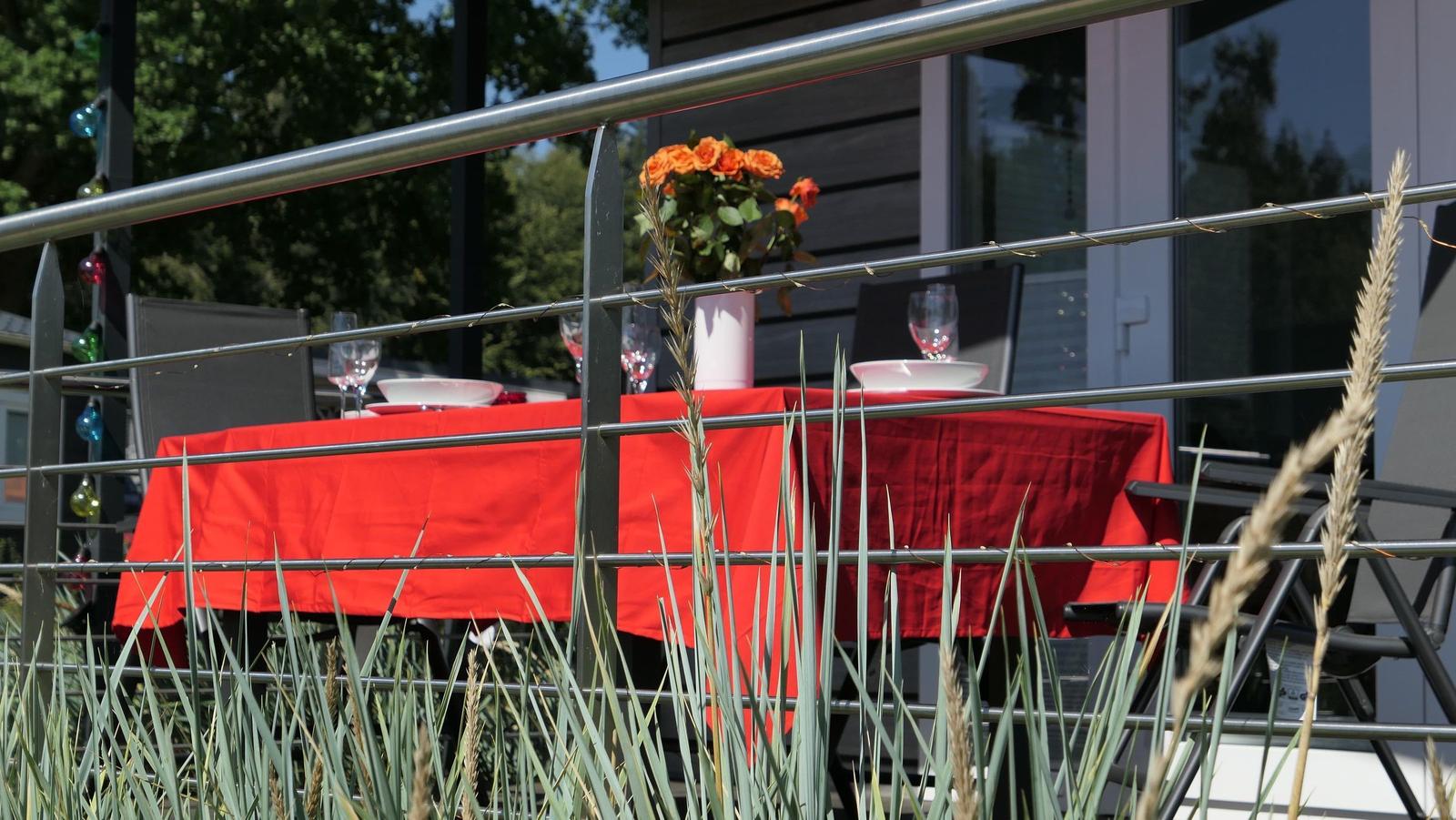 Tisch mit roter Tischdecke, Blumen und Gläser auf Balkon mit Metallgeländer.