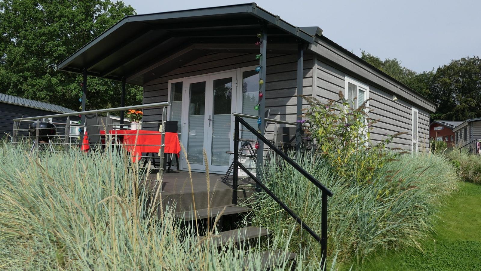 Haus mit Terrasse, Tisch, Stühlen und Grill. Umgeben von Gras und Bäumen.