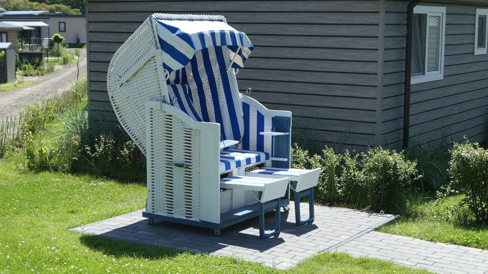 Blau-weiße Strandliege mit Schirm steht auf Steinplatten vor einem Holzhaus.
