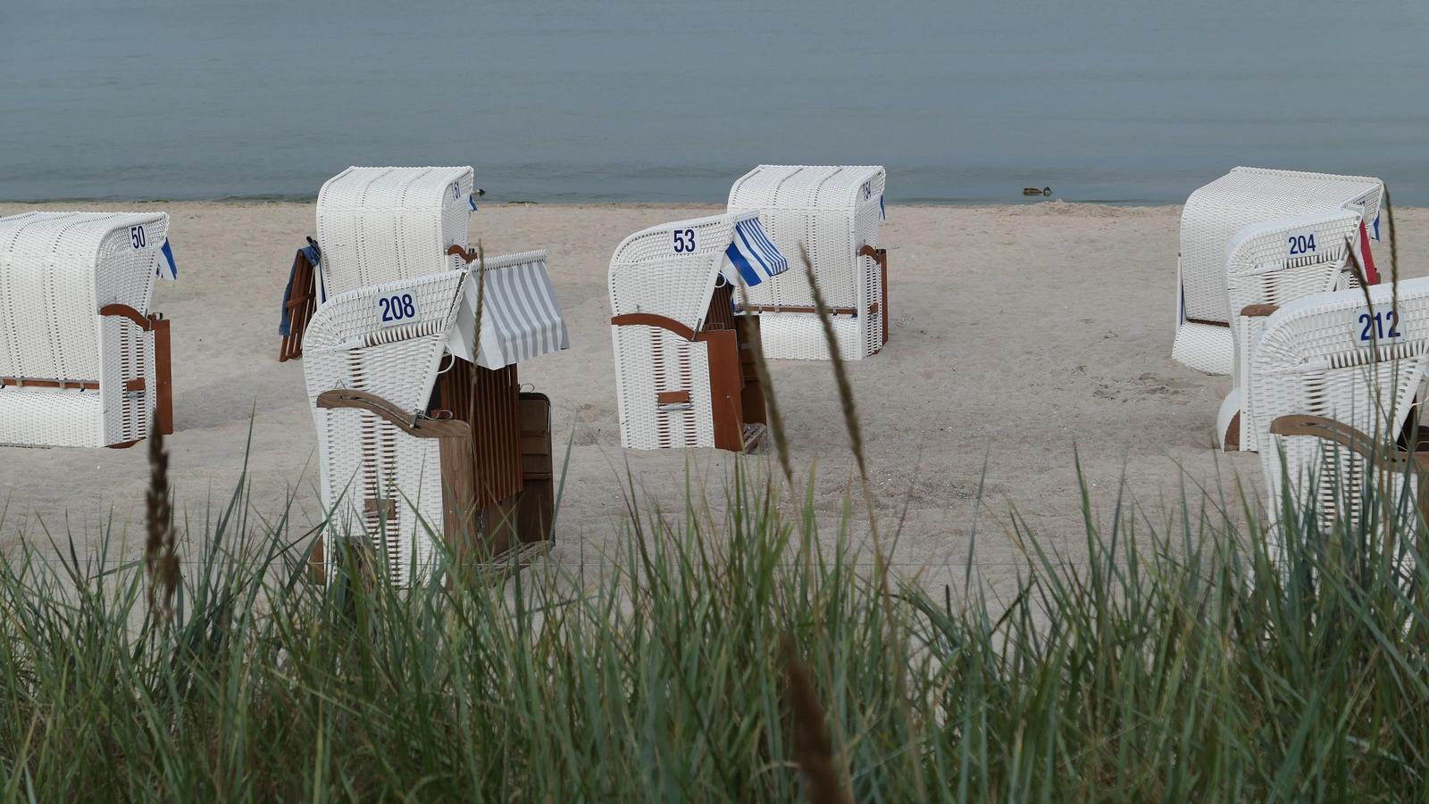 Weiße Strandkörbe mit Nummern stehen am Sandstrand vor dem Wasser.