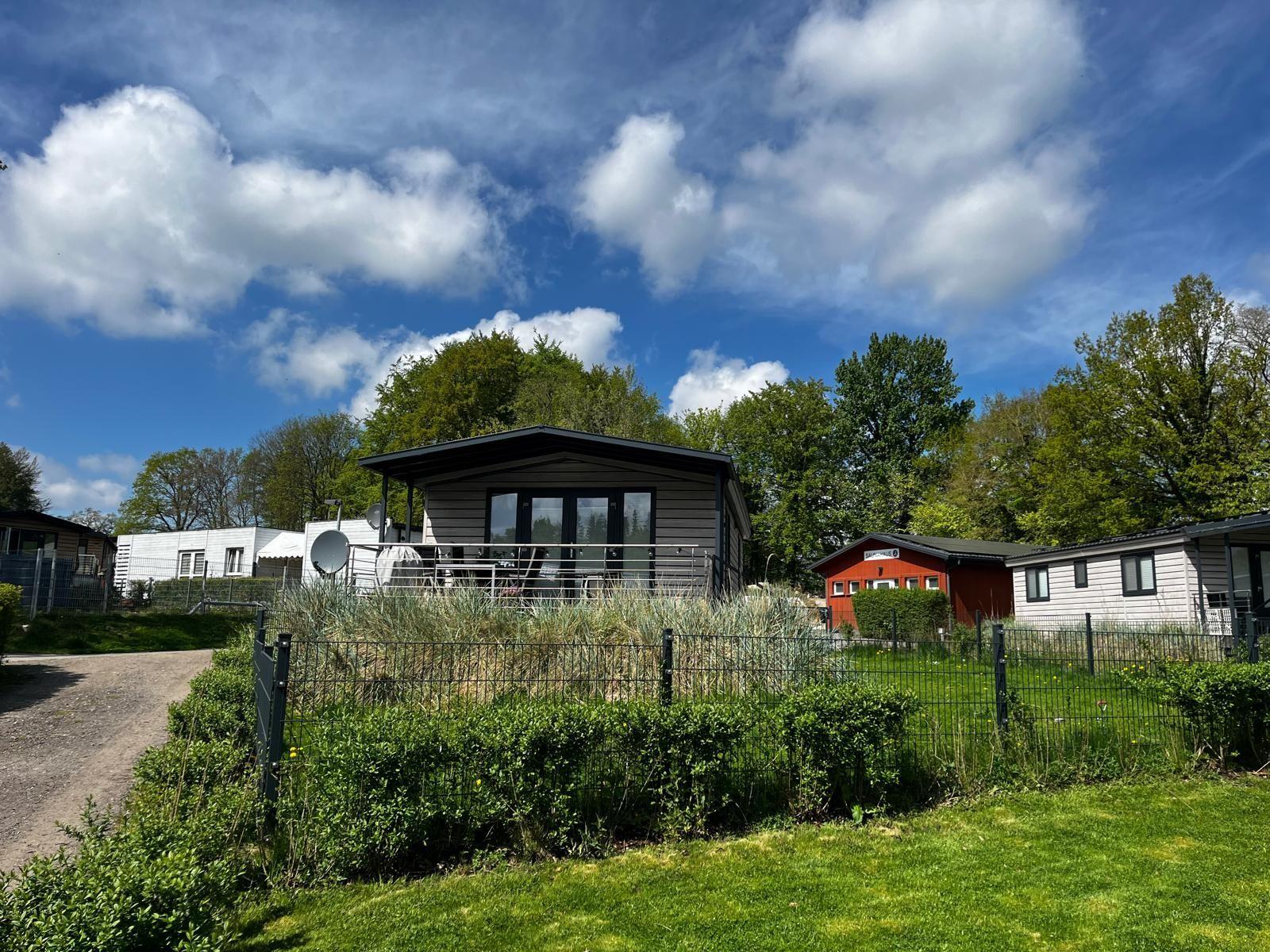 Moderne Hütte mit Terrasse, umgeben von Grün und Bäumen unter blauem Himmel.