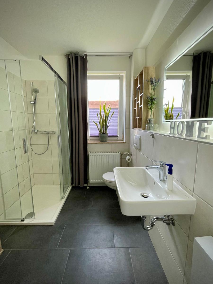 Badezimmer mit Dusche, Waschbecken und Toilette. Fenster mit Pflanze und Spiegel mit HOME.