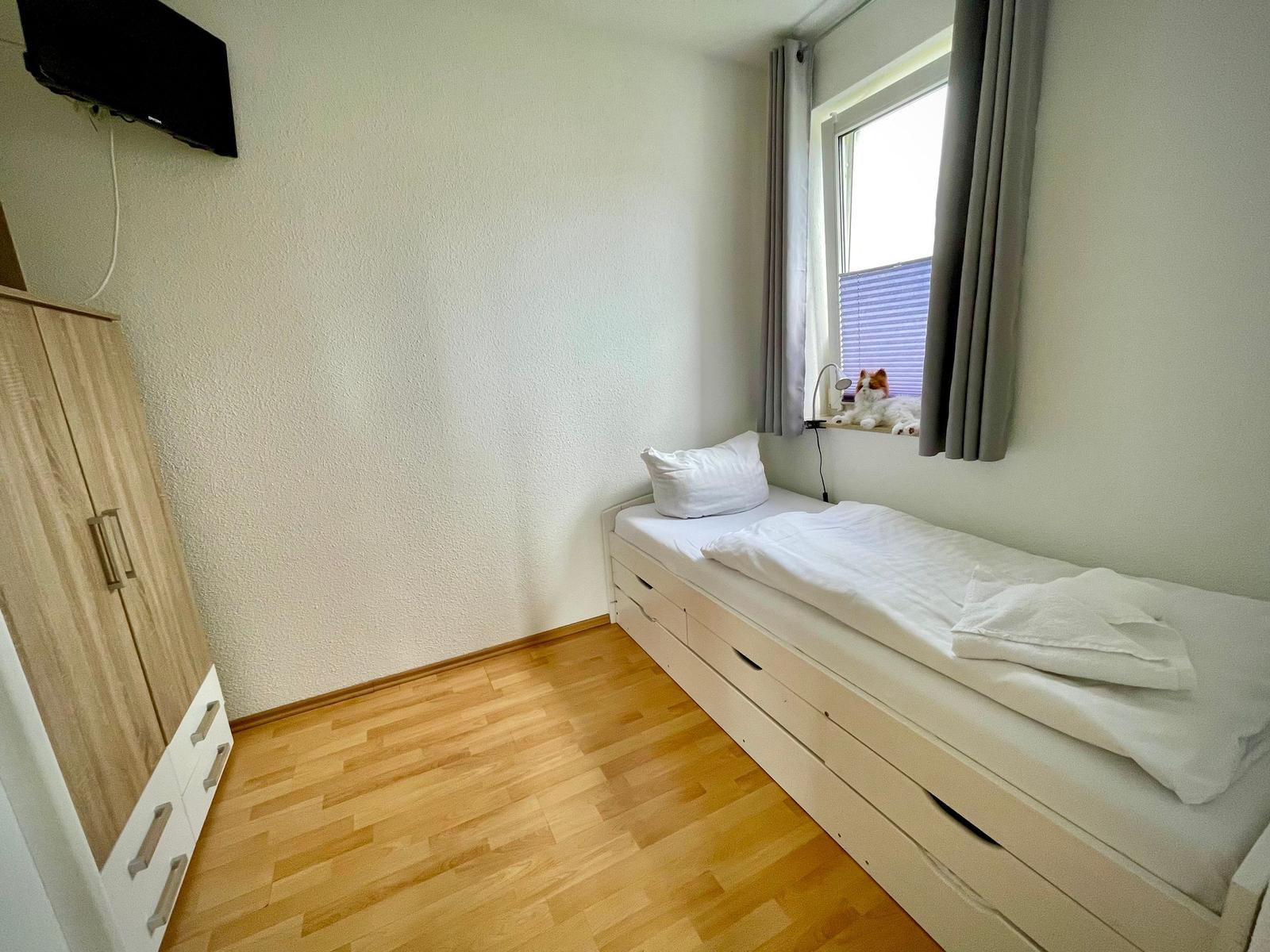 Schlafzimmer mit Bett, Kleiderschrank und Fernseher. Katze sitzt auf dem Fensterbrett.