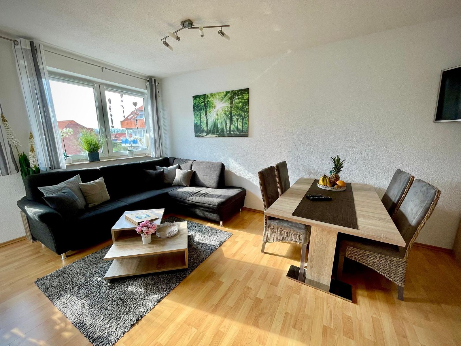 Wohn- und Essbereich mit Sofa, Tisch und Stühlen. Fenster mit Blick auf Außenbereich.