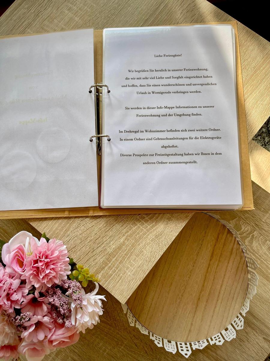 Info-Broschüre mit Hinweisen zur Ferienwohnung und Umgebung liegt auf Holztisch mit Blumen.