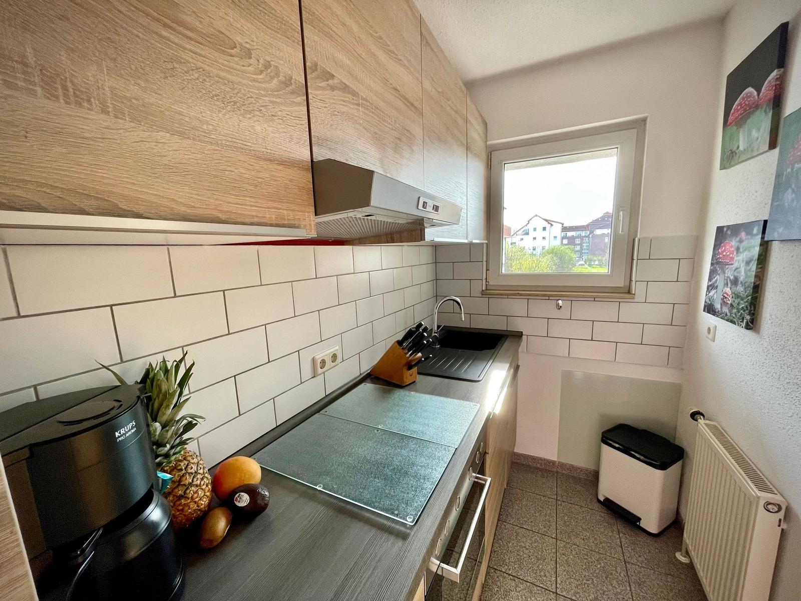 Küche mit Holzschränken, Spüle und Fenster. Auf der Arbeitsfläche stehen Obst und eine Kaffeemaschine.