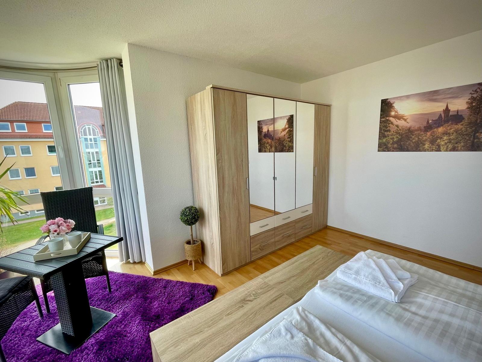 Schlafzimmer mit Doppelbett, Kleiderschrank und Balkon mit Sitzgelegenheit.