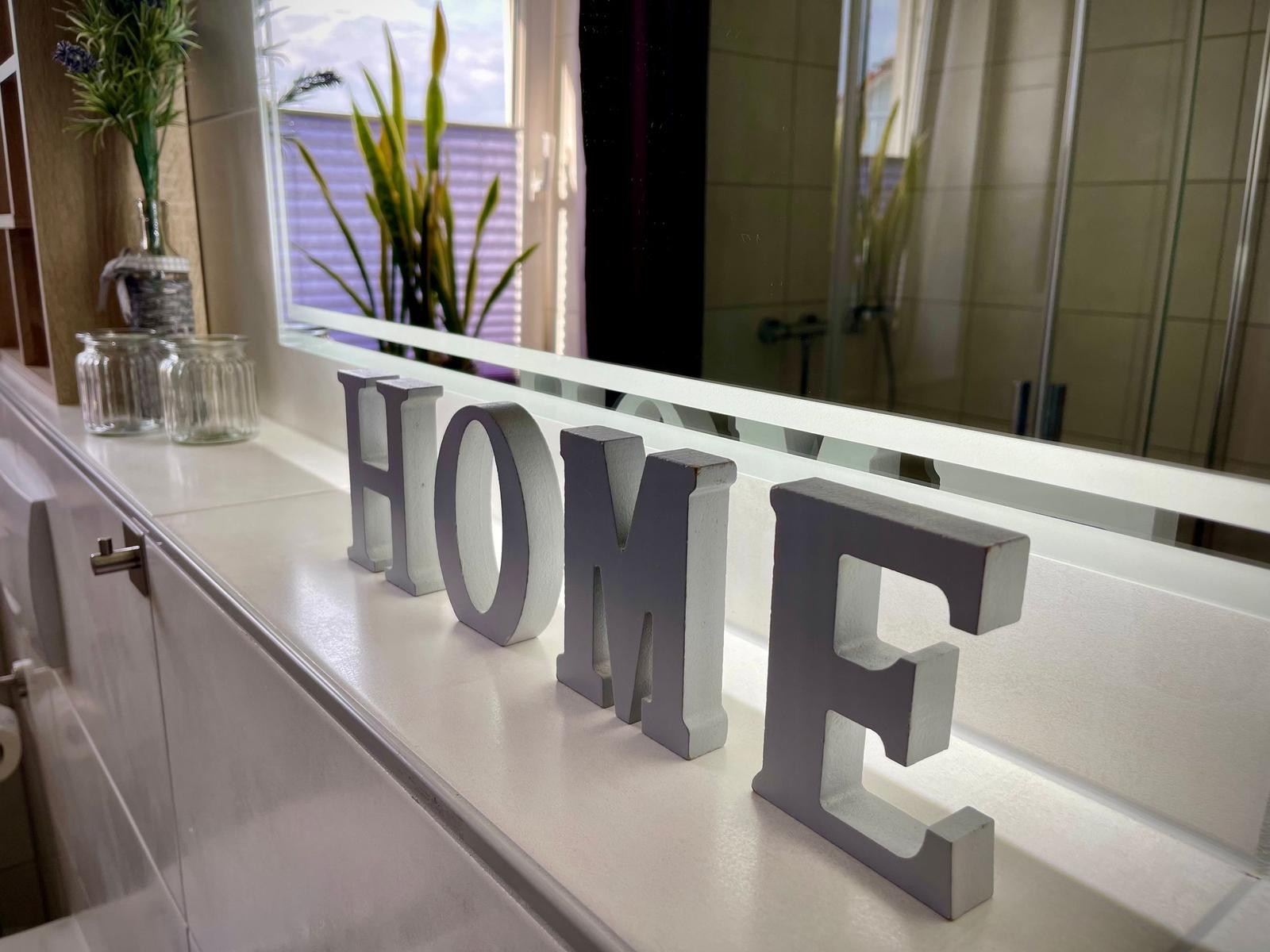 Badezimmer mit Spiegel, Pflanzen und grauen Buchstaben HOME auf der Arbeitsplatte.