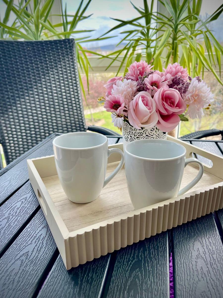 Zwei weiße Tassen auf einem Holzbrett mit Blumenarrangement im Hintergrund.