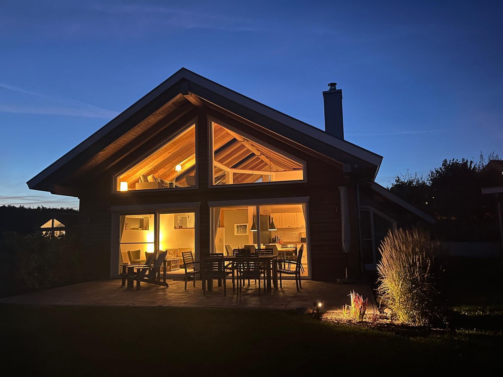 Haus mit großem Holzrahmen, beleuchteten Fenstern und Terrasse mit Tisch und Stühlen.