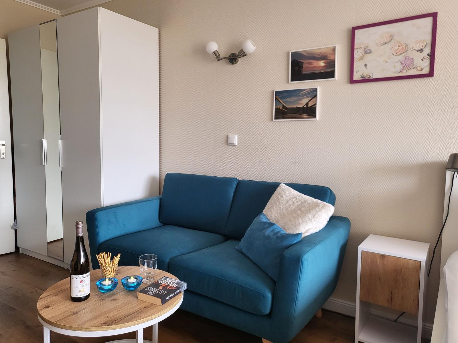 Blue sofa with cushions, round table with wine and book, wall art and wardrobe.