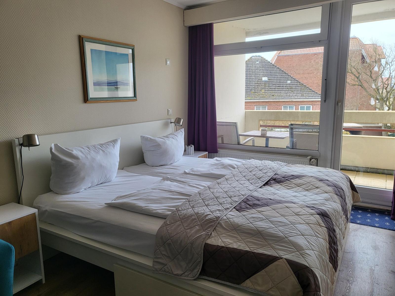Double bed with white bedding and quilt. Balcony with table and chairs. Wall art and nightstand.
