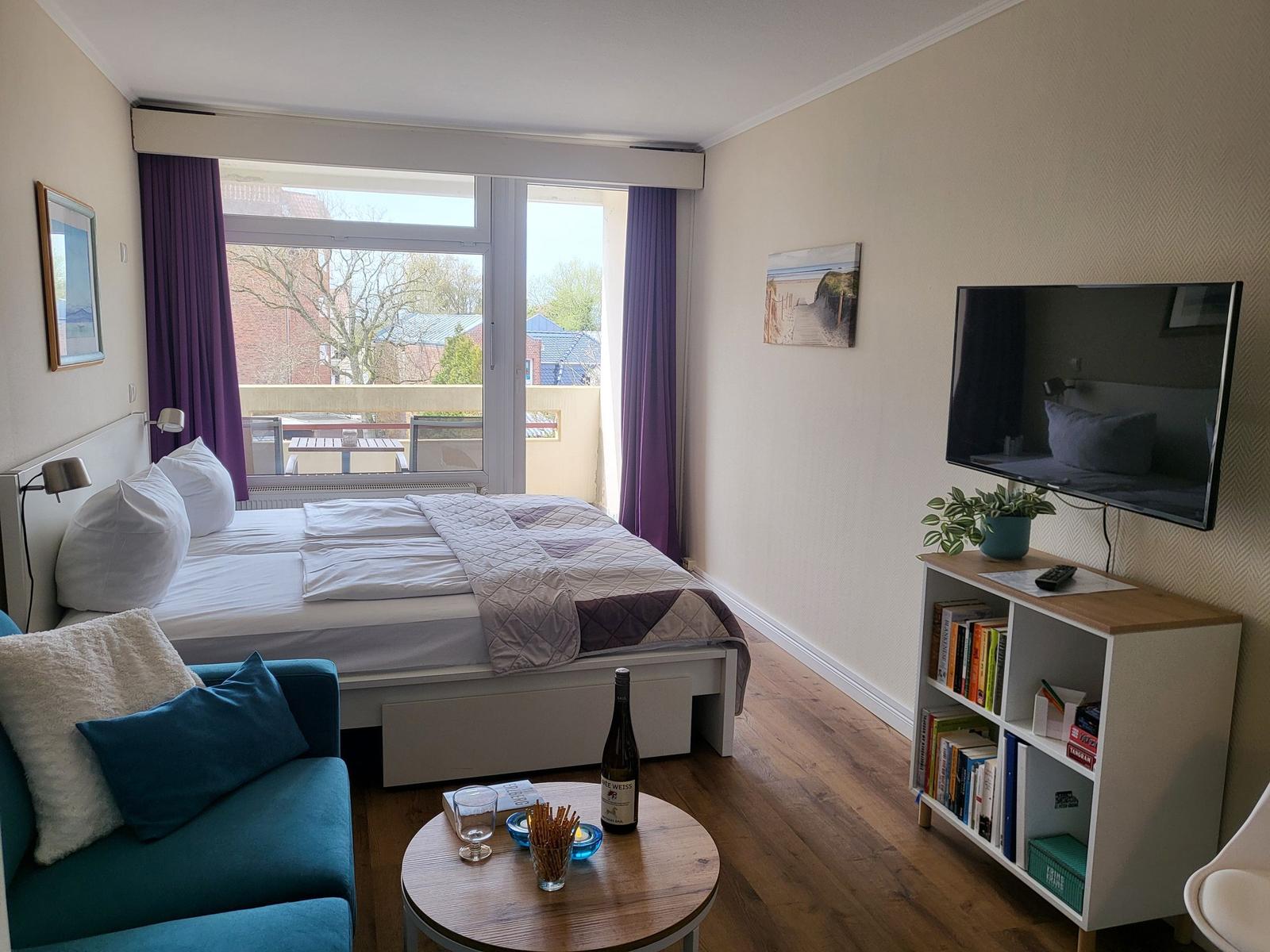Bedroom with bed, TV, and balcony. Wooden floor and bookshelf.