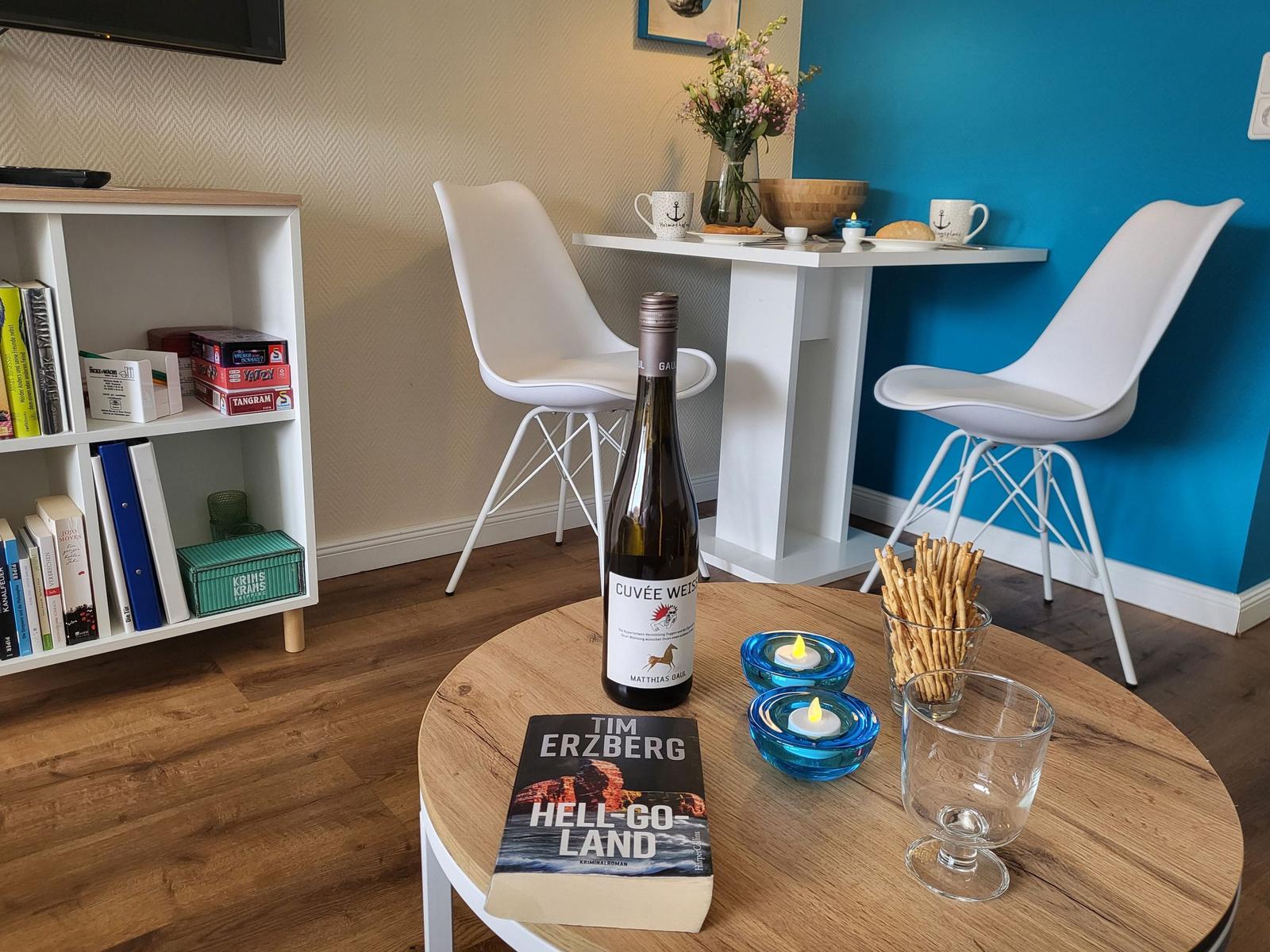 Round table with wine, book, and candles. Background: dining table with chairs and bookcase.