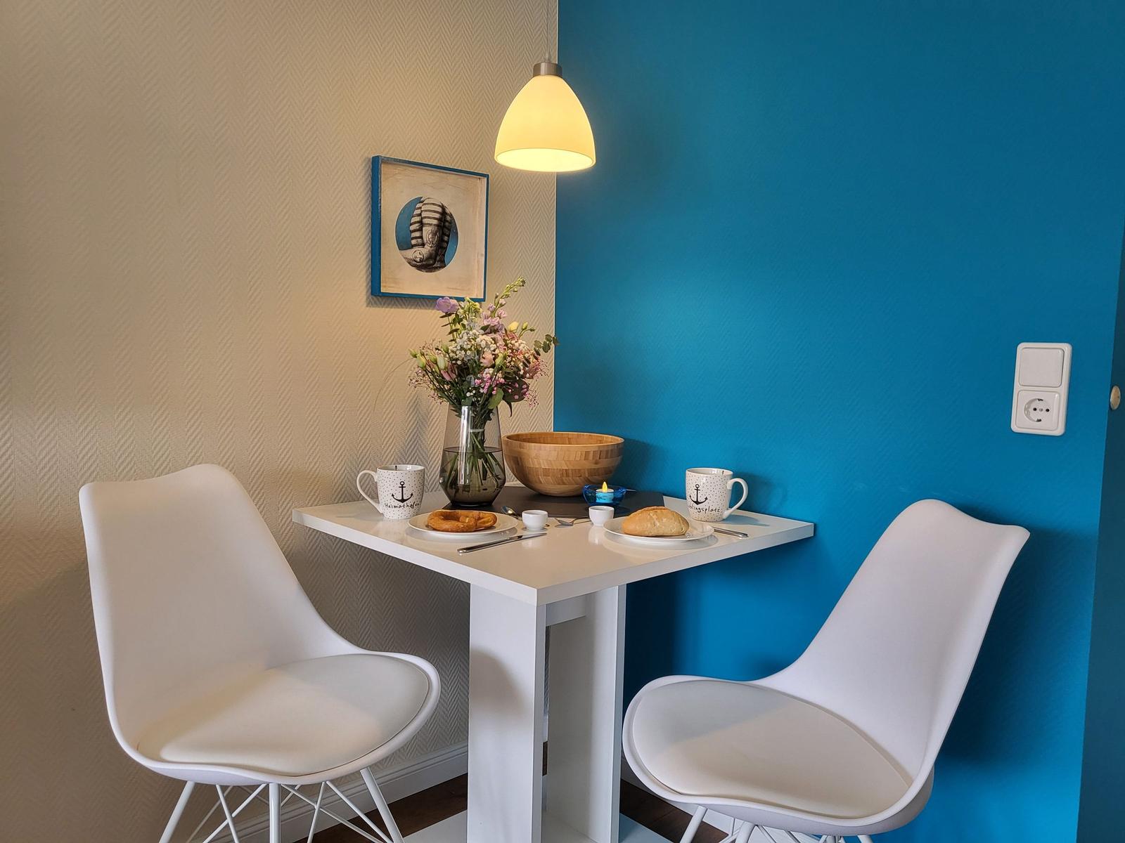 Dining table with two chairs, flowers, and coffee mugs against blue wall.