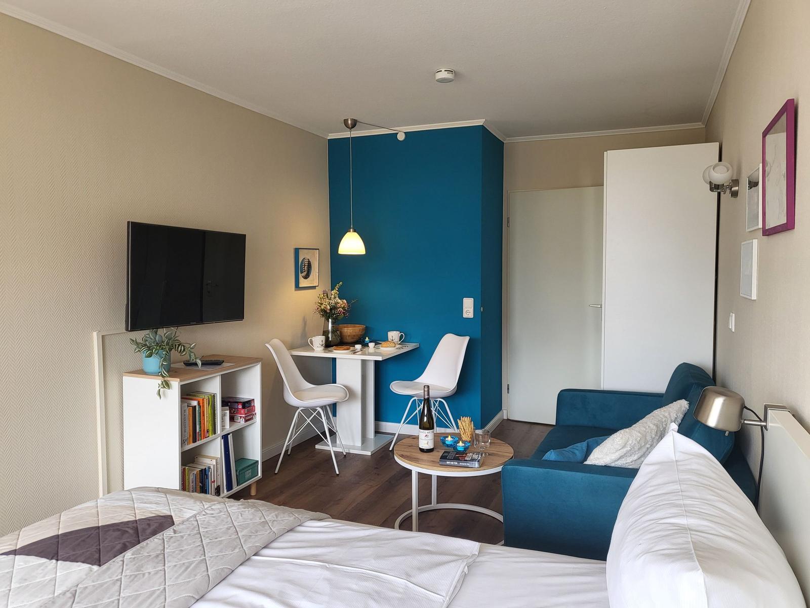 Bedroom with sofa, dining area, and TV. Blue wall, wooden floor, and bookshelf.