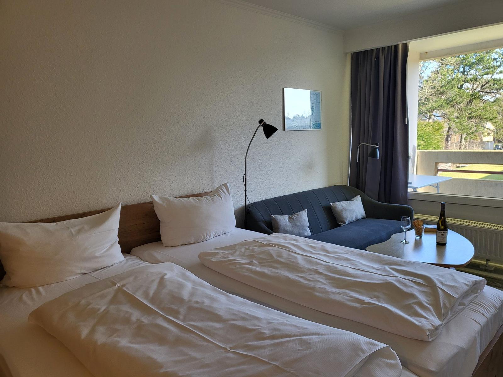 Zwei Betten, Sofa und Tisch mit Weinflasche und Glas vor Fenster mit Blick auf Grünfläche.