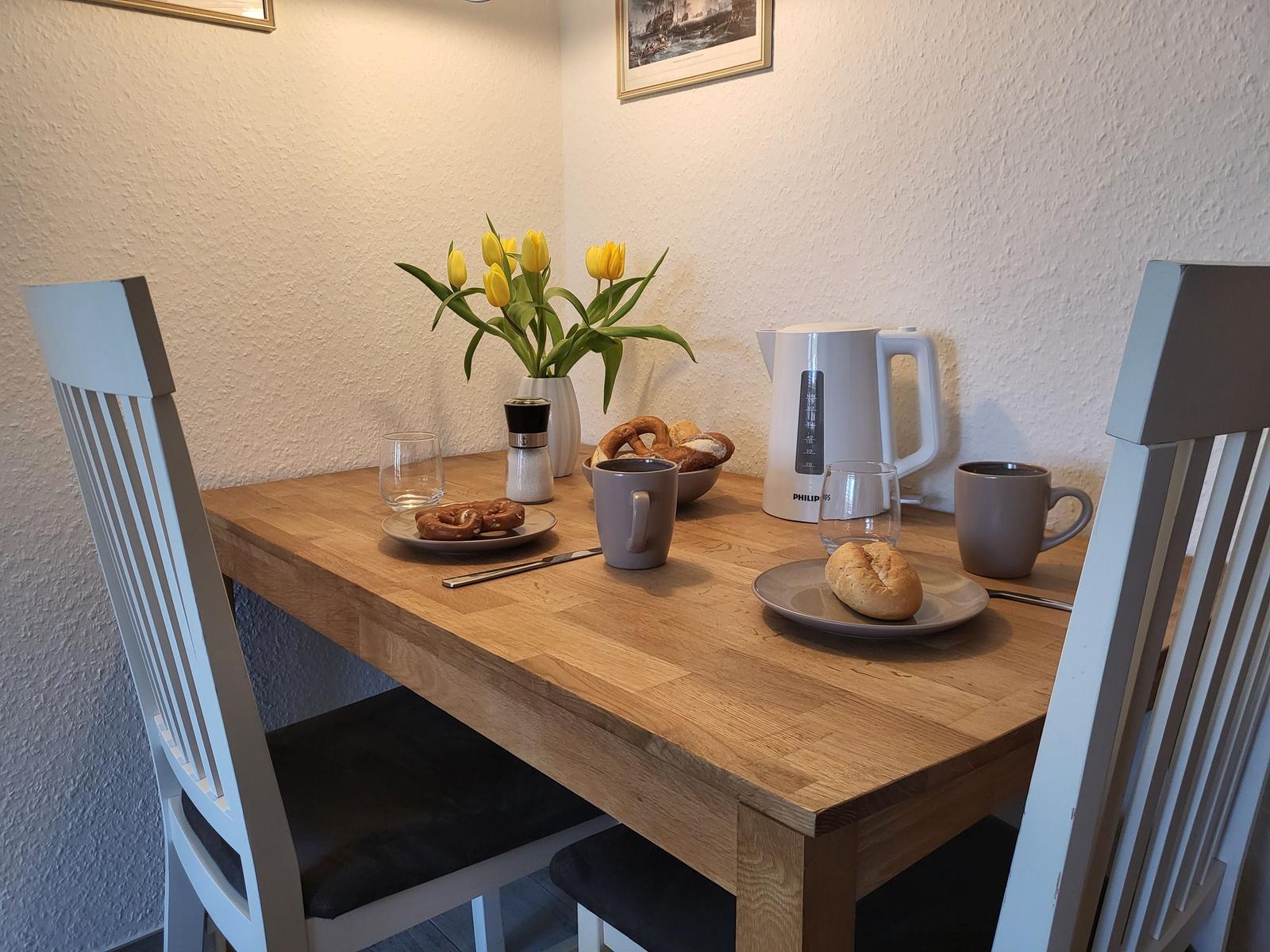Holztisch mit Stühlen, Kaffeekanne, Brötchen und Tulpenblumen.