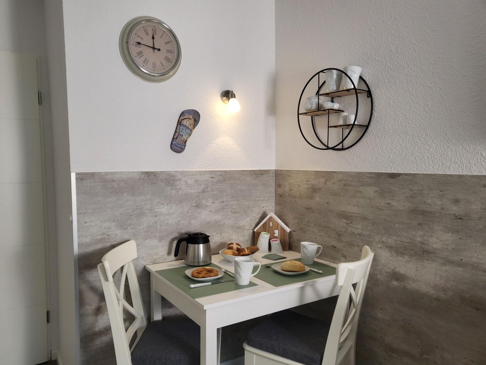 Esstisch mit zwei Stühlen, Kaffeekanne und Brötchen. Wanduhr und Regal mit Tassen.