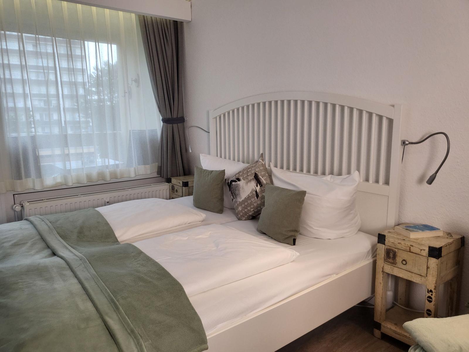 Ein Bett mit weißen Laken und grünen Kissen. Neben dem Bett steht ein Nachttisch.