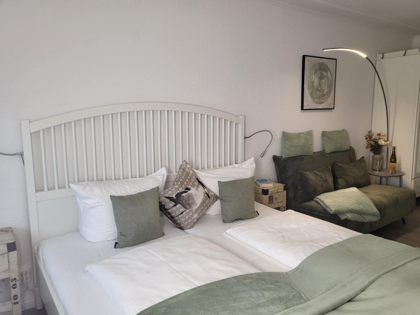 Schlafzimmer mit Doppelbett, grünen Kissen und Sofa. Wandbild und Stehlampe.