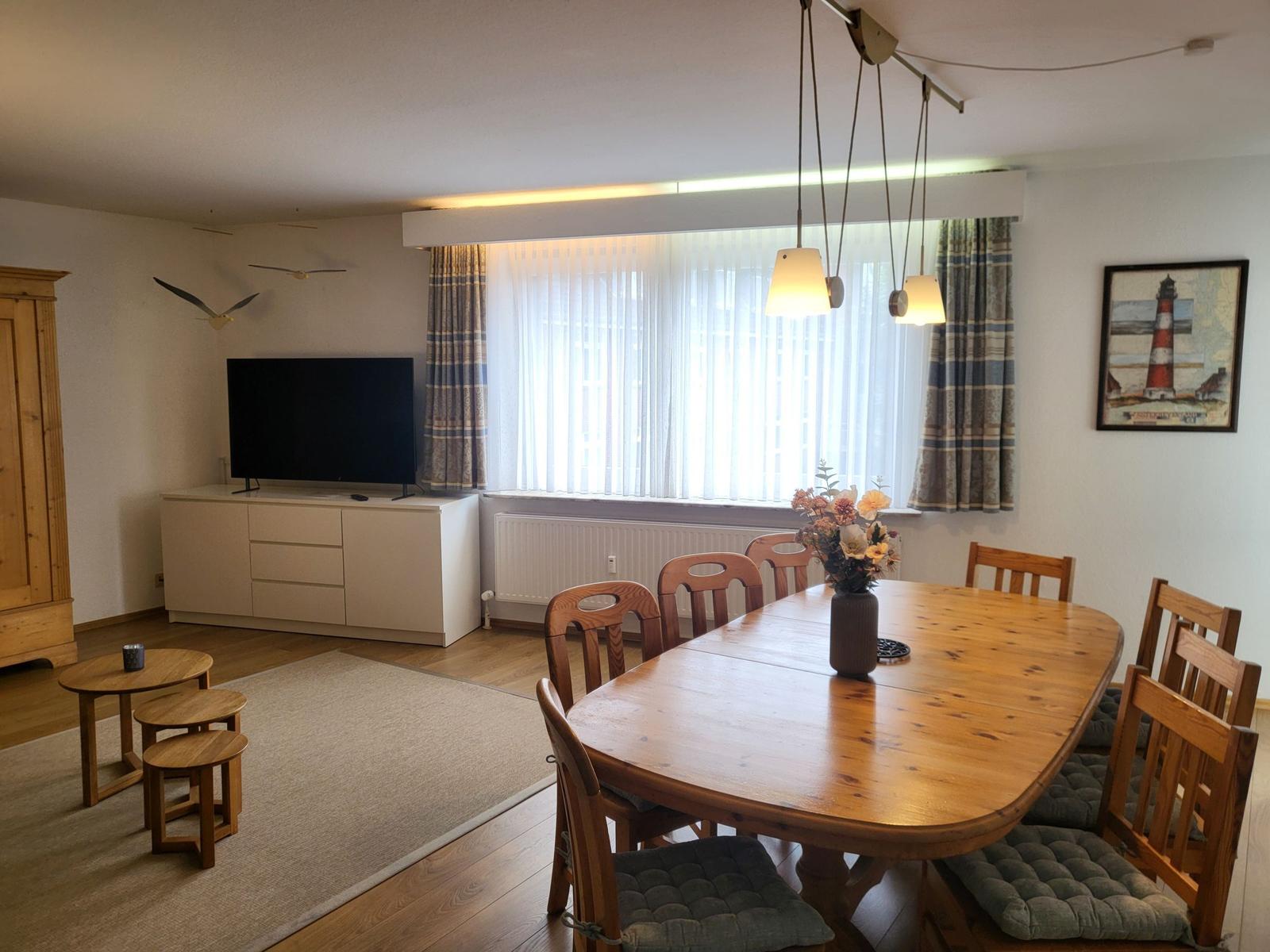 Essbereich mit Holztisch und Stühlen, Fernseher und Fenster mit Vorhängen.