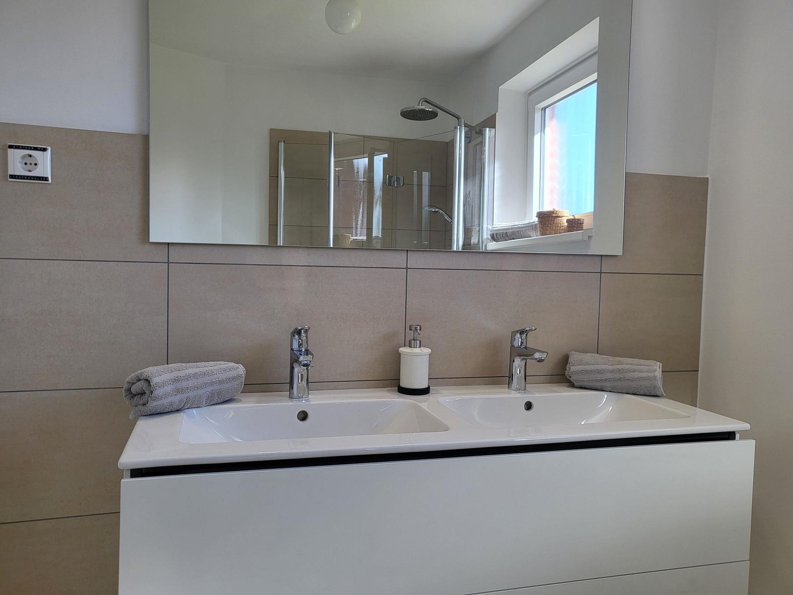 Bathroom with double sink, mirror, and shower. Window with view.