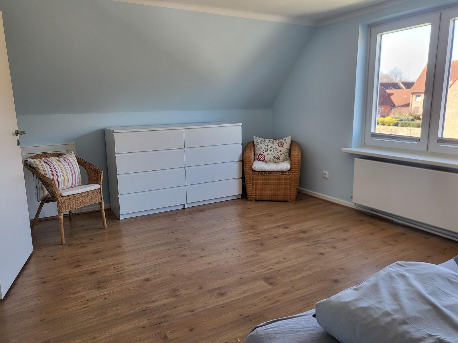 A bedroom with wooden floor, white dresser, and two chairs by a window.