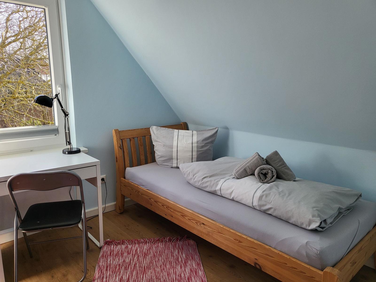 A bedroom with wooden bed, desk and window. The walls are light blue.