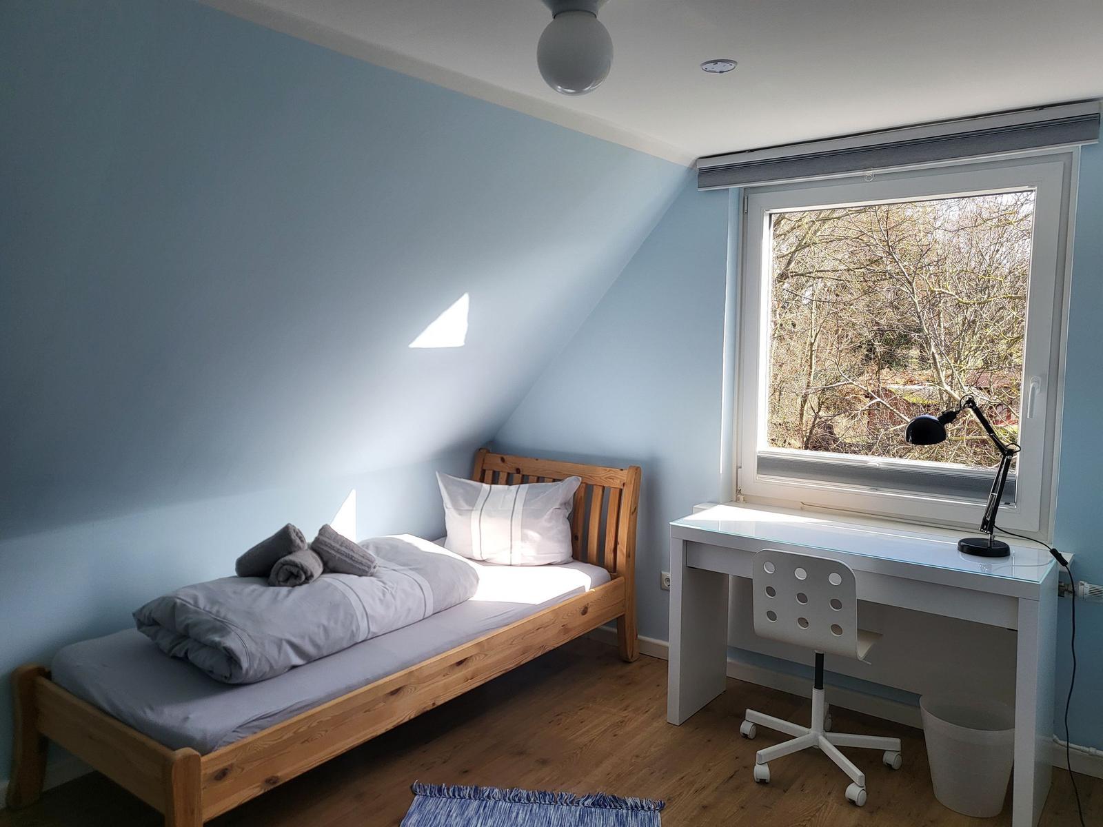 A bedroom with sloped ceiling, bed, desk, and window with view of trees.